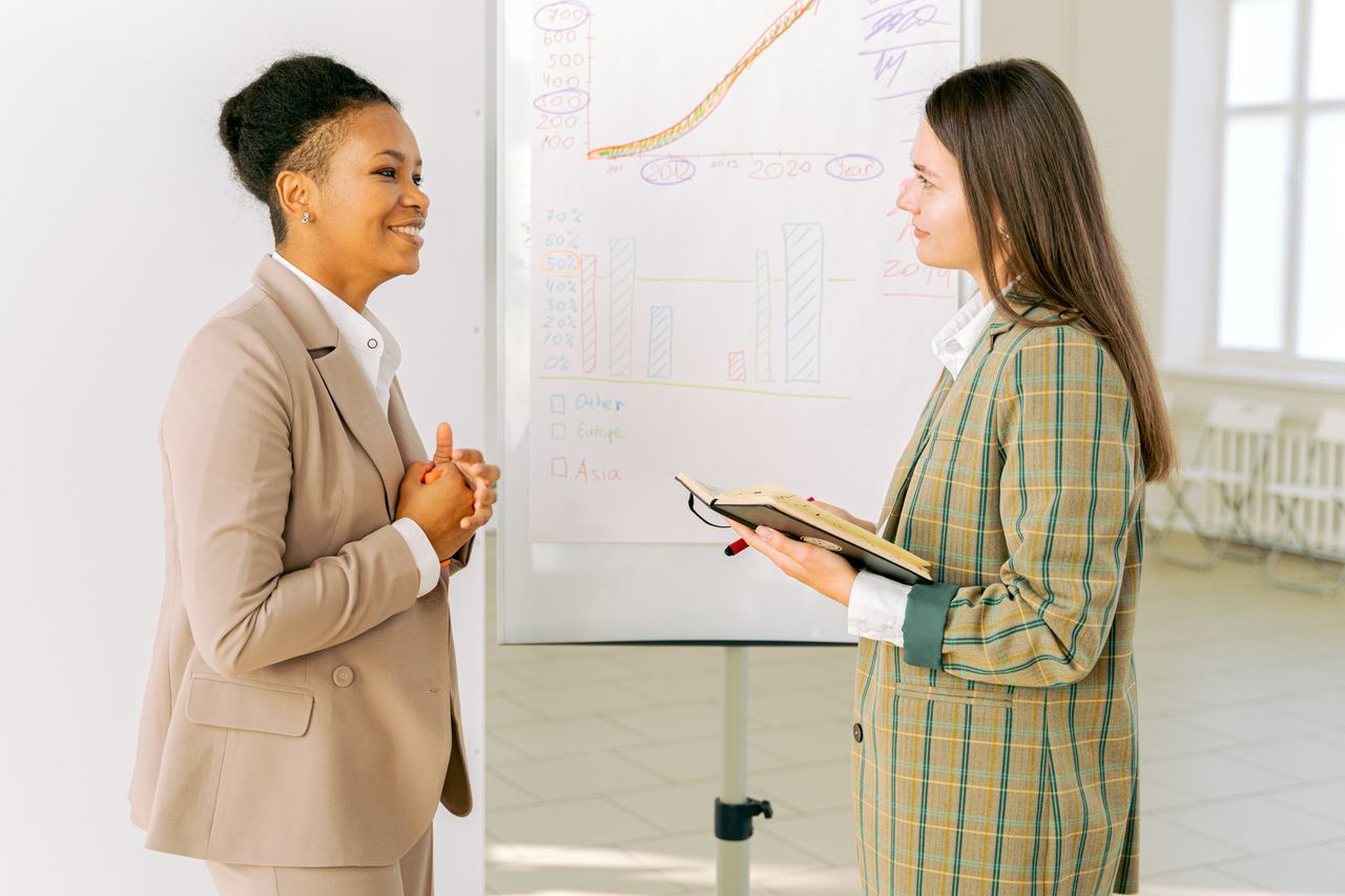Businesswomen discussing strategies by a whiteboard in a modern office setting.